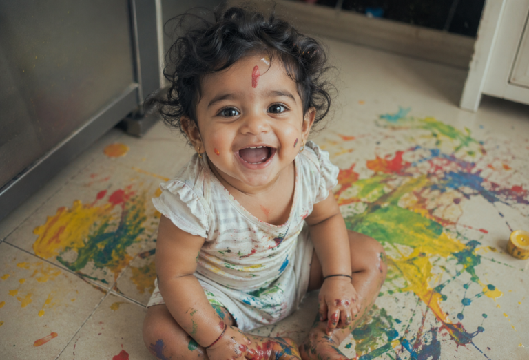 baby laughing while sitting on kitchen floor covered in colorful paint during a messy art session