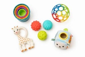 Best toys for 6 month old baby flat lay including stacking cups OBall textured balls Sophie the Giraffe and soft fill and spill taco toy on white background