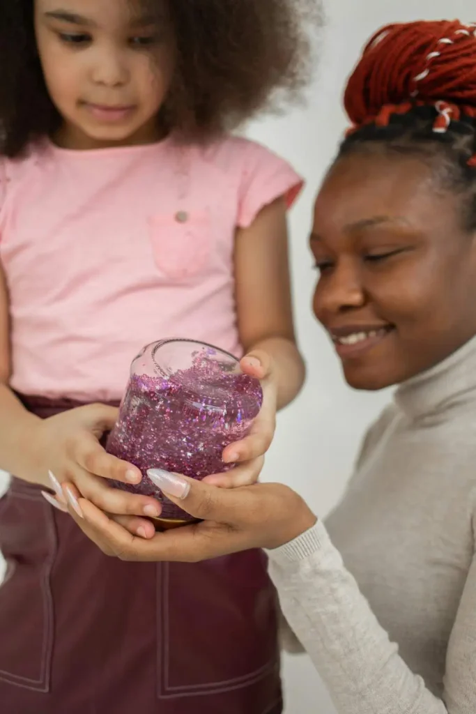 A toddler’s hands holding a clear plastic glitter calm sensory bottle filled with blue water and slow‑falling silver glitter, designed to help kids focus, calm down, and regulate big emotions during meltdowns or transitions.
