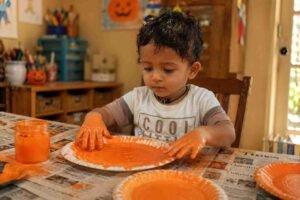 Preschool child painting a paper plate orange for a Halloween pumpkin craft with paint-covered hands and a small orange paint spot on the nose.