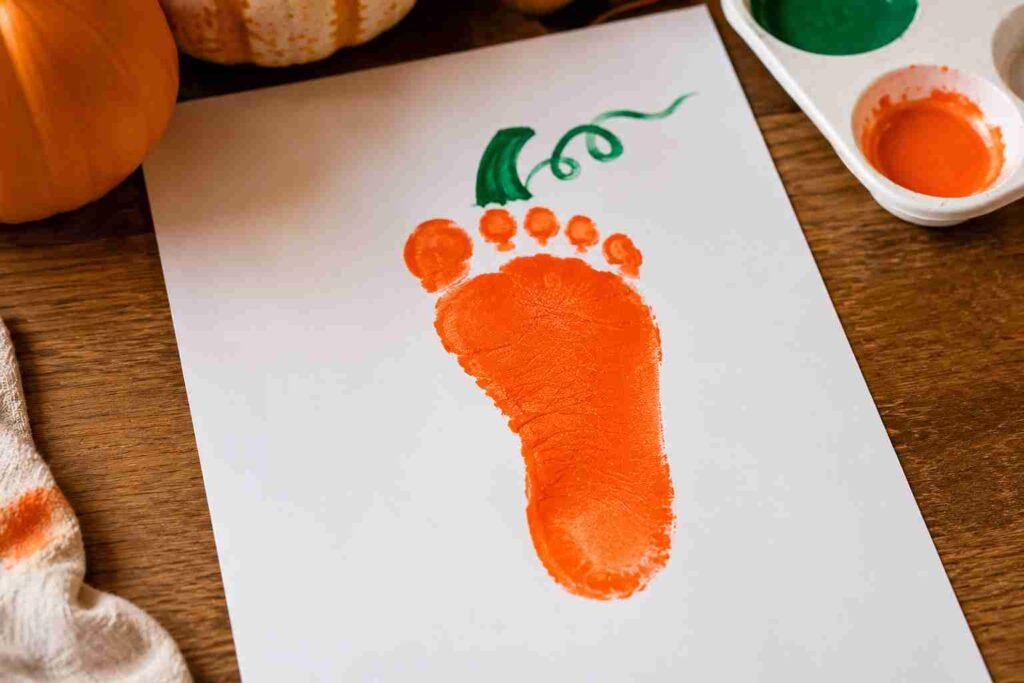 Orange baby footprint pumpkin craft on white paper with a green stem, creating an easy Halloween keepsake for babies and toddlers.