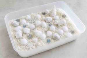 A white winter sensory bin with rice, cotton balls, and a wooden spoon for babies and toddlers