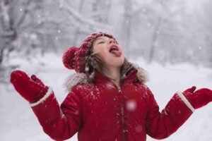 A child in a red coat and mittens catching snowflakes with their mouth open in a snowy garden